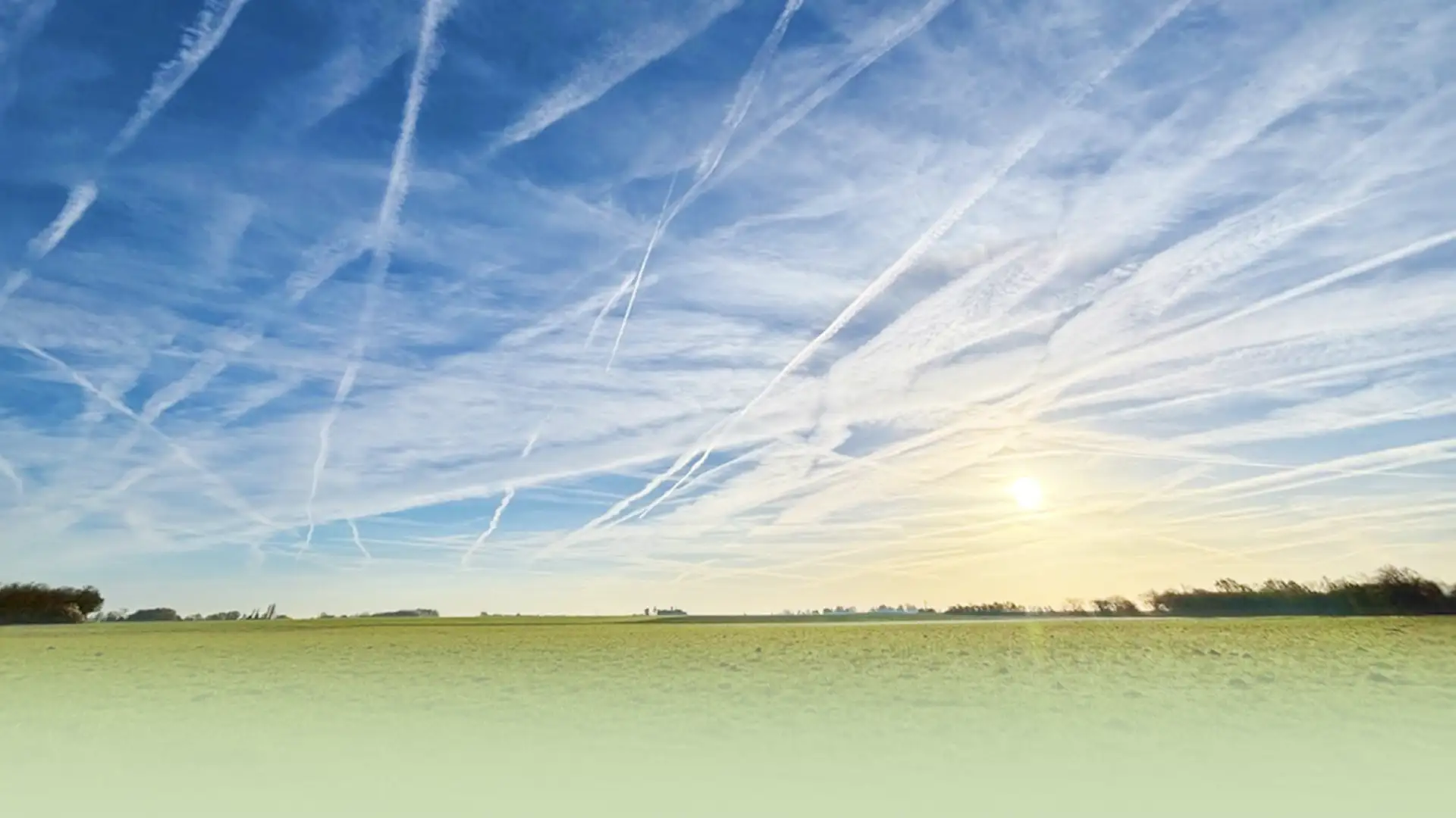 Von zahlreichen Kondens- und Aerosolstreifen durchzogener Himmel über einer Landschaft als Symbol für Geoengineering, Wettermanipulation und großflächige Eingriffe in die Atmosphäre.