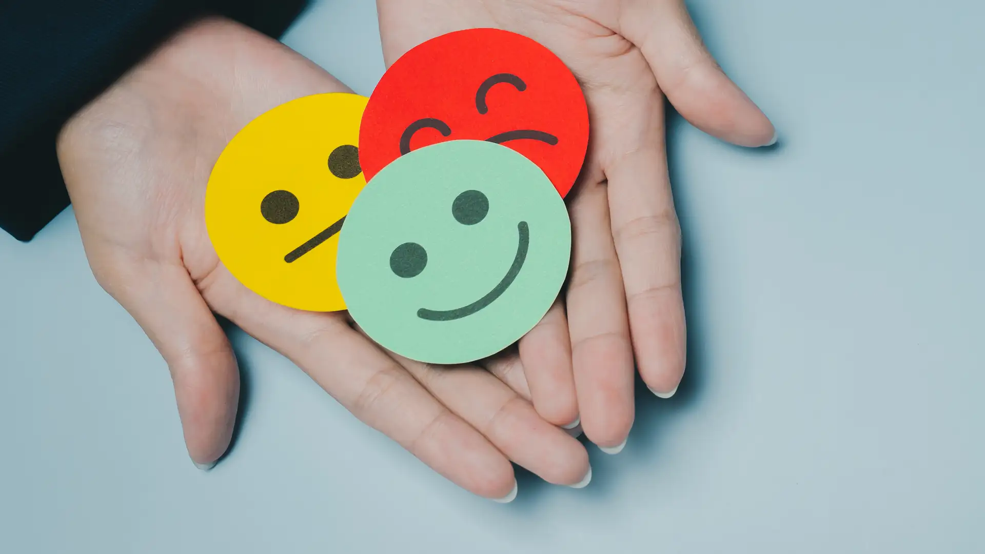 Hände halten drei verschiedenfarbige Emotionssymbole mit fröhlichem, neutralem und traurig-frustriertem Gesicht.